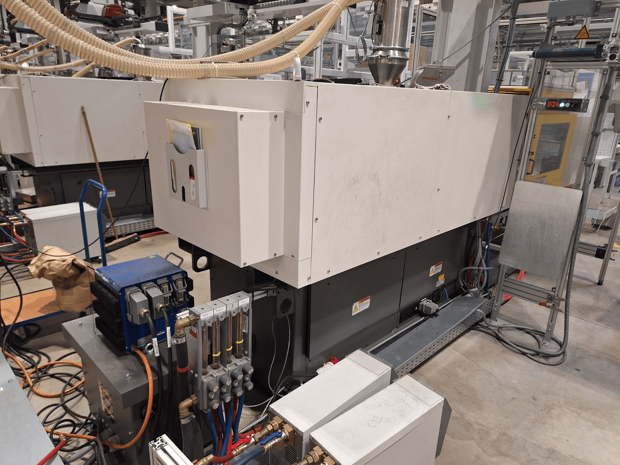 Pramoninė įranga gamykloje; didelė mašina su valdymo skydeliu, žarnomis ir elektros jungtimis, vaizdas iš šono.