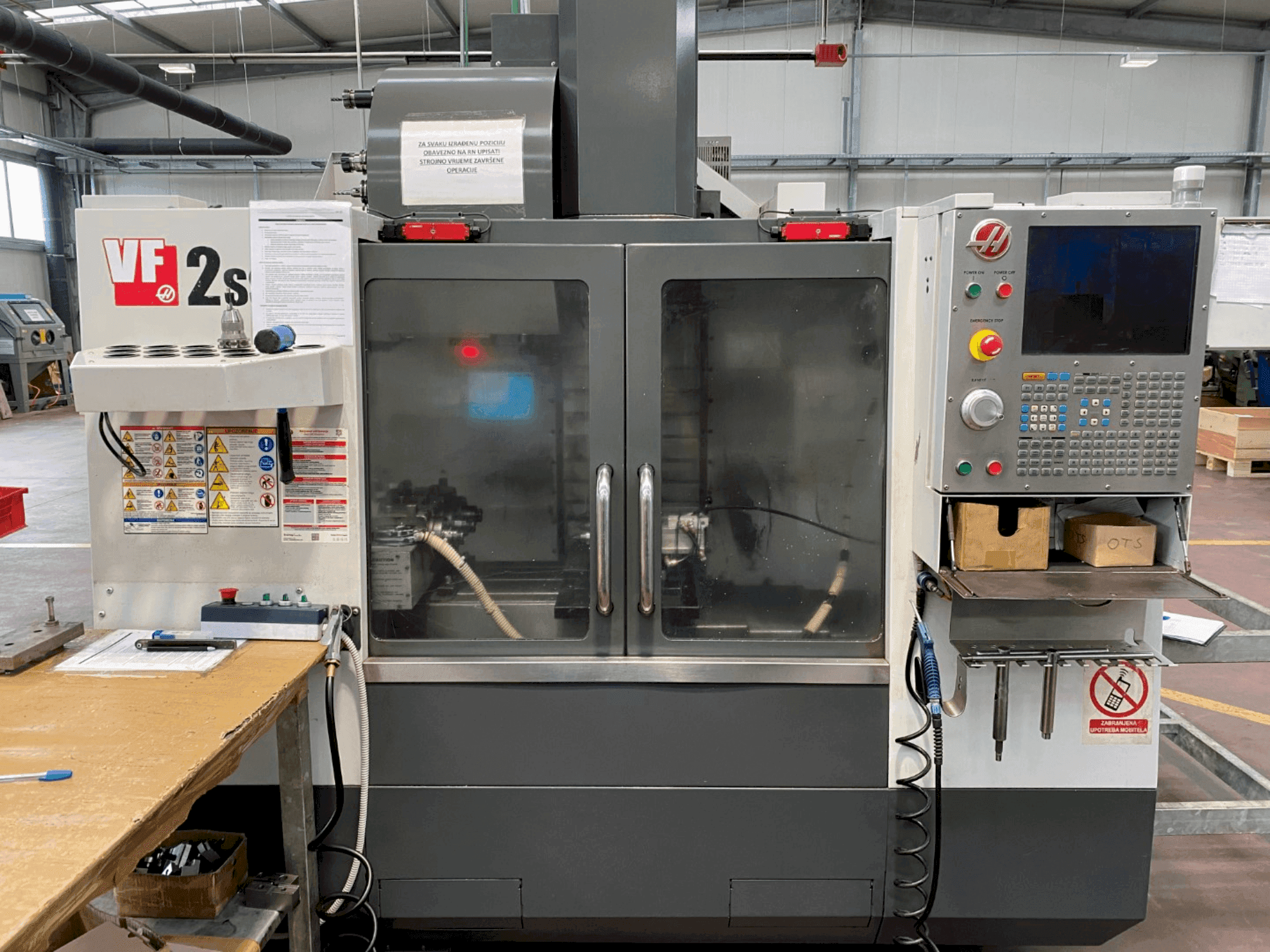 "Haas VF-2SS" apdirbimo centras iš priekio, su valdymo skydeliu, aušinimo skysčio žarna ir saugos etiketėmis šone.
