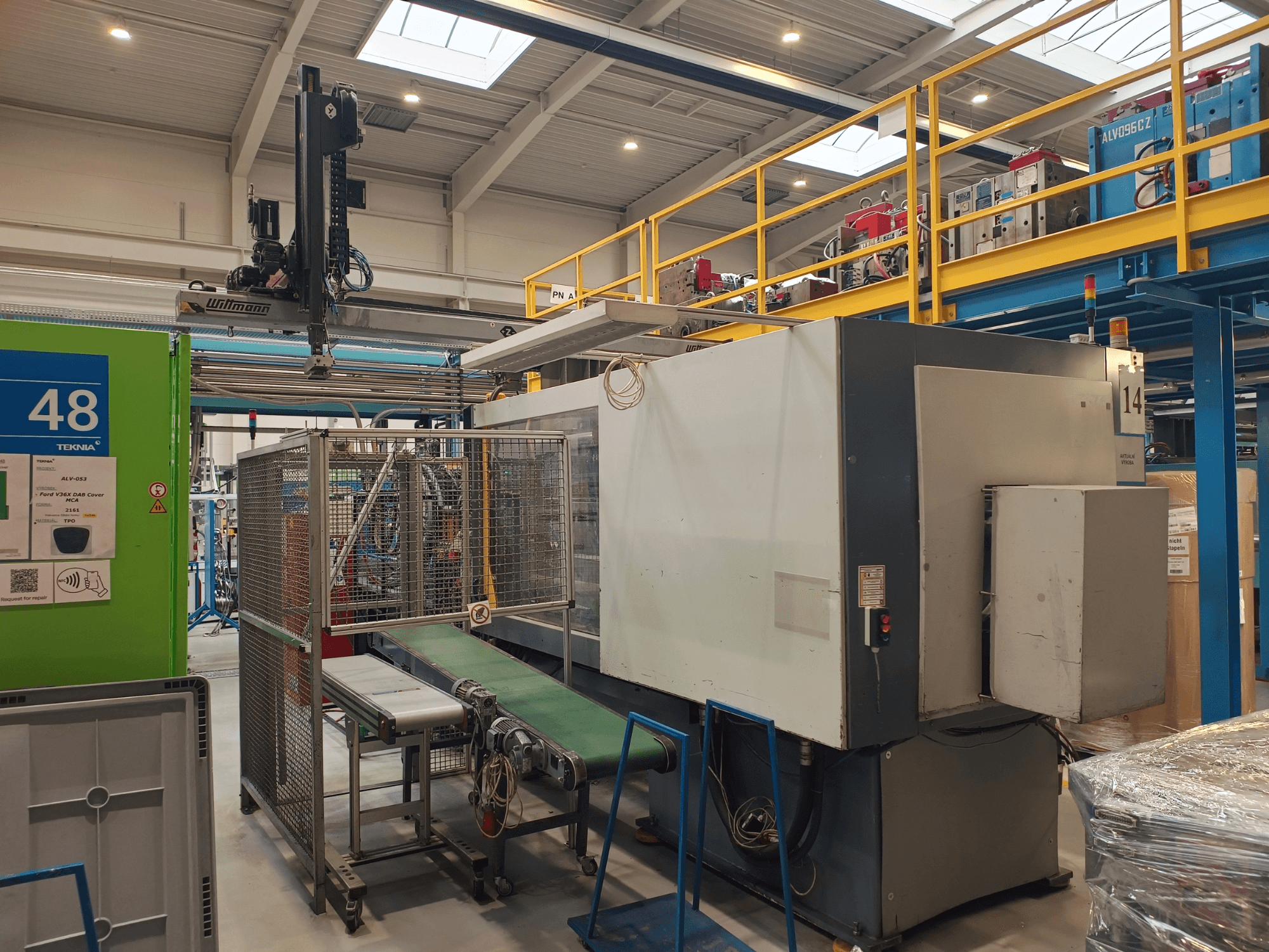 Pramonės automatizavimo įranga su žaliu konvejeriu, pažymėtu "48", ir tiltiniu kranu; mašinos vaizdas iš šono ant gamyklos grindų.